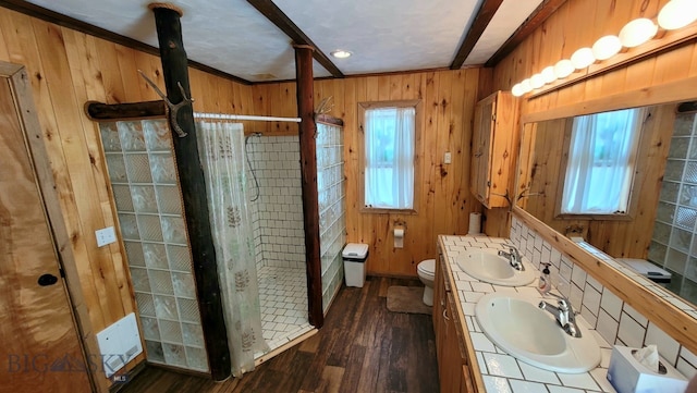 bathroom with wooden walls, double vanity, tiled shower, toilet, and wood-type flooring