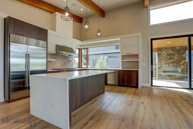 kitchen featuring appliances with stainless steel finishes, hanging light fixtures, dark brown cabinets, wall chimney range hood, and beam ceiling