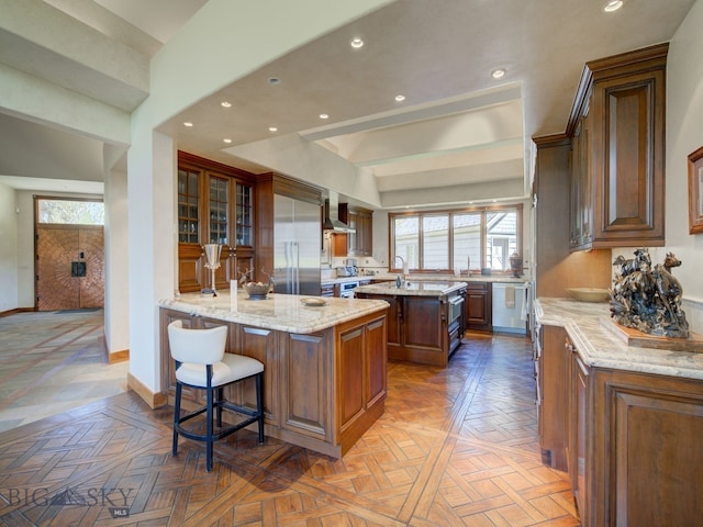 kitchen featuring parquet floors, wall chimney range hood, an island with sink, light stone counters, and a breakfast bar