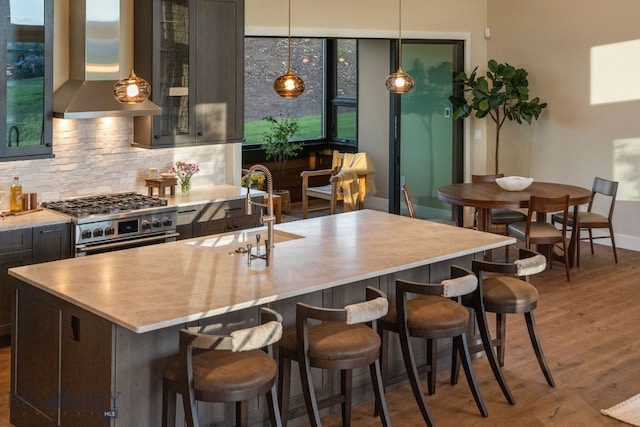 kitchen featuring wood-type flooring, wall chimney exhaust hood, sink, stainless steel stove, and a center island with sink