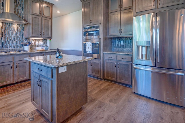 kitchen with wall chimney range hood, stainless steel appliances, wood-type flooring, light stone countertops, and a kitchen island