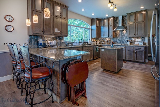 kitchen featuring wall chimney exhaust hood, hanging light fixtures, dark stone countertops, a kitchen island, and decorative backsplash