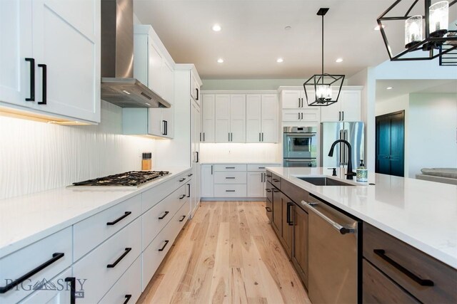 kitchen featuring light wood-type flooring, wall chimney range hood, white cabinets, stainless steel appliances, and pendant lighting