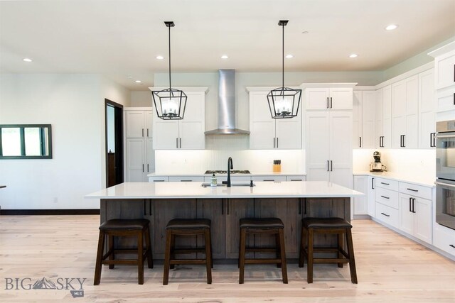 kitchen featuring a kitchen island with sink, white cabinetry, wall chimney exhaust hood, and light wood-type flooring