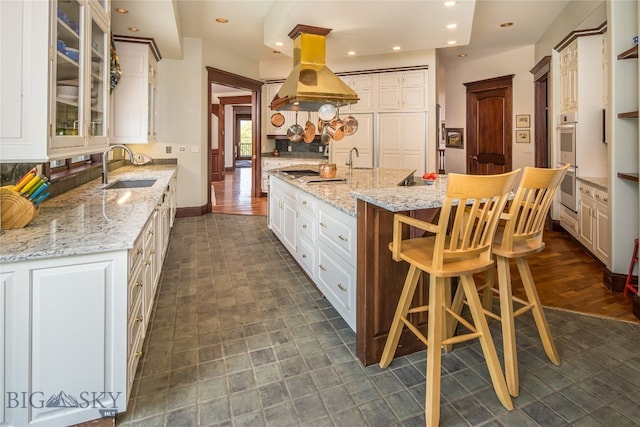 kitchen featuring island range hood, light stone countertops, sink, an island with sink, and white cabinets