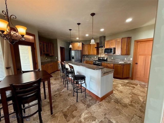 kitchen with stone tile floors, stainless steel appliances, dark countertops, wall chimney exhaust hood, and a notable chandelier