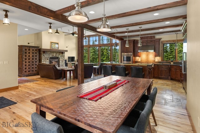 dining room with ceiling fan, beam ceiling, a stone fireplace, and light wood-type flooring