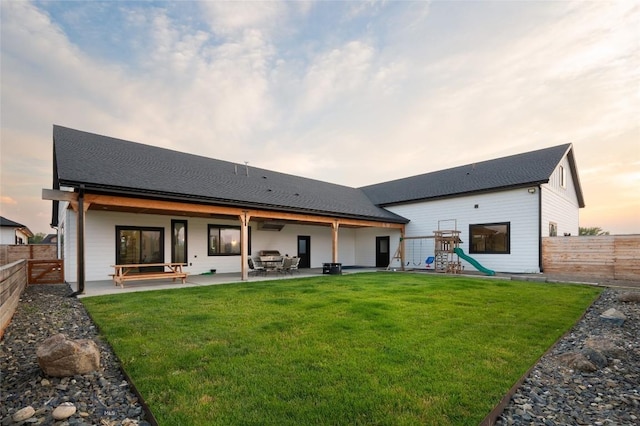 back house at dusk with a playground, a patio area, and a lawn