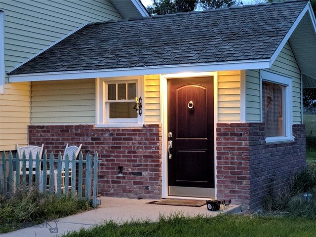 entrance to property featuring a shingled roof, brick siding, and fence