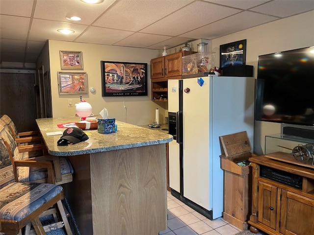 kitchen with light tile patterned flooring, white refrigerator with ice dispenser, light stone counters, a kitchen bar, and a drop ceiling