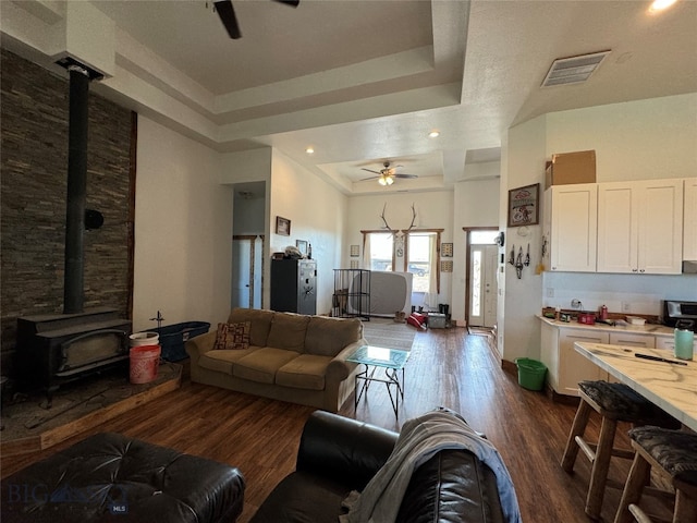 living room with dark hardwood / wood-style flooring, a tray ceiling, a wood stove, and ceiling fan