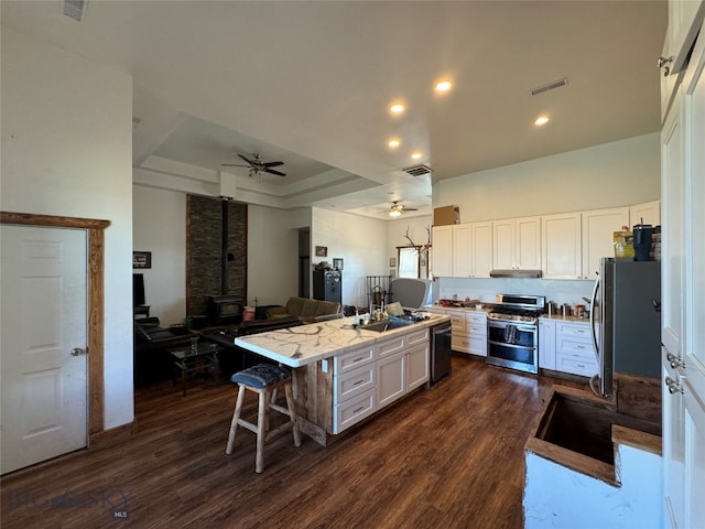 kitchen with stainless steel appliances, a tray ceiling, a center island with sink, white cabinets, and a wood stove