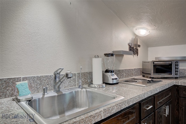 kitchen with a textured ceiling, vaulted ceiling, dark brown cabinets, sink, and stainless steel cooktop