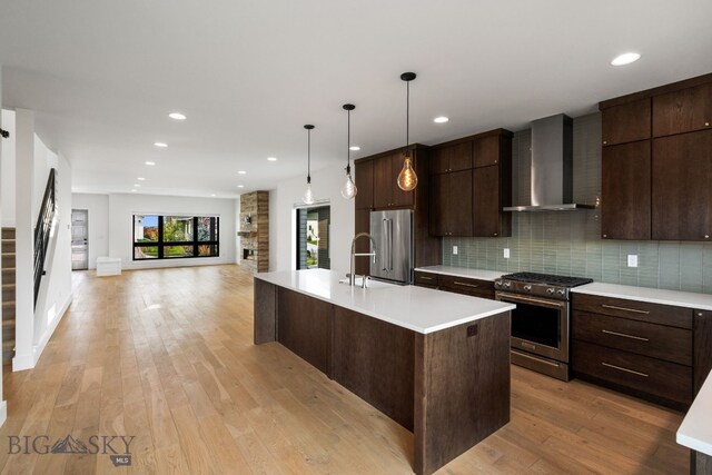 kitchen featuring high end appliances, wall chimney exhaust hood, decorative light fixtures, a center island with sink, and backsplash