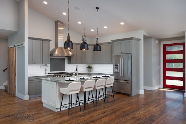 kitchen featuring dark hardwood / wood-style floors, wall chimney exhaust hood, a kitchen island with sink, stainless steel appliances, and light stone counters