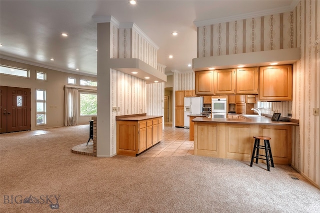 kitchen featuring kitchen peninsula, white appliances, light colored carpet, a breakfast bar area, and crown molding