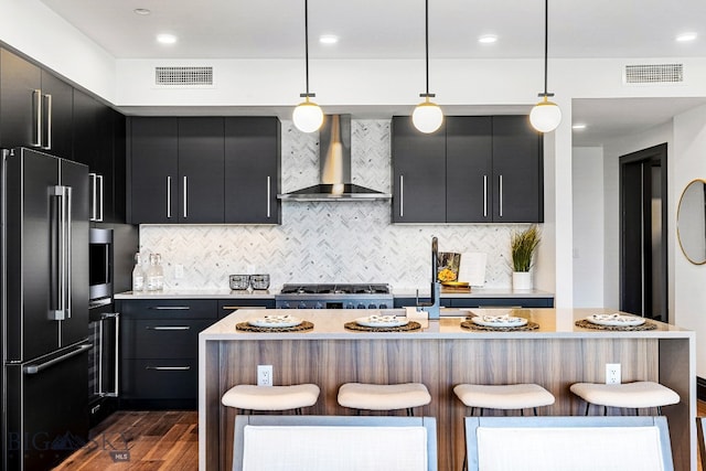 kitchen with wall chimney range hood, a center island with sink, a kitchen breakfast bar, pendant lighting, and stainless steel appliances