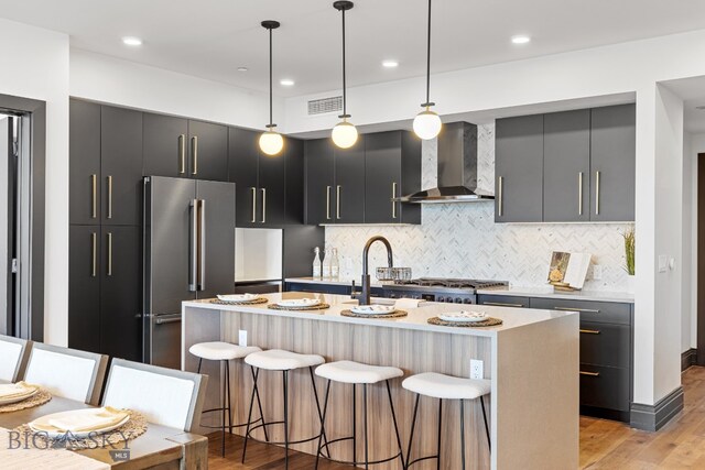 kitchen with appliances with stainless steel finishes, wall chimney exhaust hood, a kitchen bar, decorative light fixtures, and a kitchen island with sink