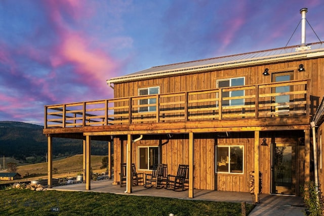 back house at dusk featuring a wooden deck, a lawn, and a patio area