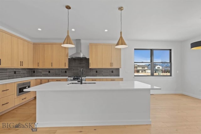 kitchen featuring light brown cabinetry, a kitchen island with sink, wall chimney range hood, pendant lighting, and light hardwood / wood-style flooring