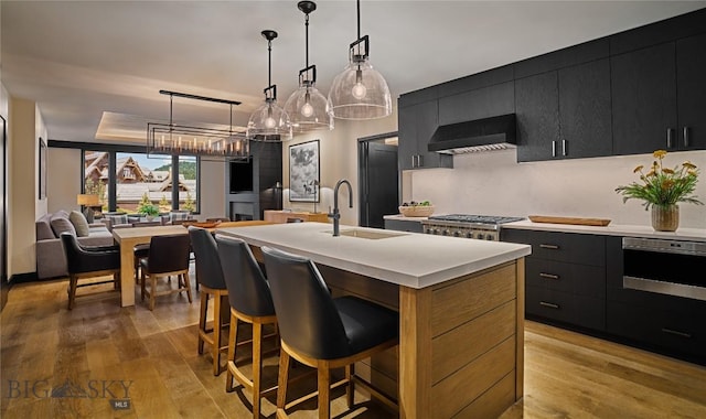 kitchen with a kitchen island with sink, a sink, hanging light fixtures, dark cabinetry, and wall chimney exhaust hood