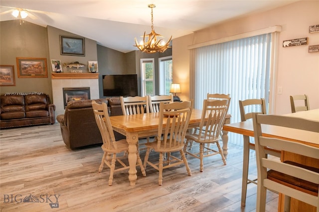 dining room with ceiling fan with notable chandelier, vaulted ceiling, light wood-type flooring, and a fireplace