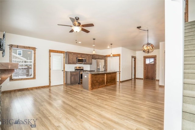 kitchen with light wood-type flooring, appliances with stainless steel finishes, a kitchen island with sink, pendant lighting, and ceiling fan with notable chandelier