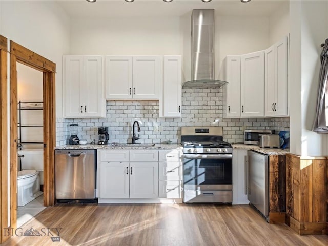 kitchen with a sink, wall chimney range hood, light stone counters, white cabinets, and appliances with stainless steel finishes