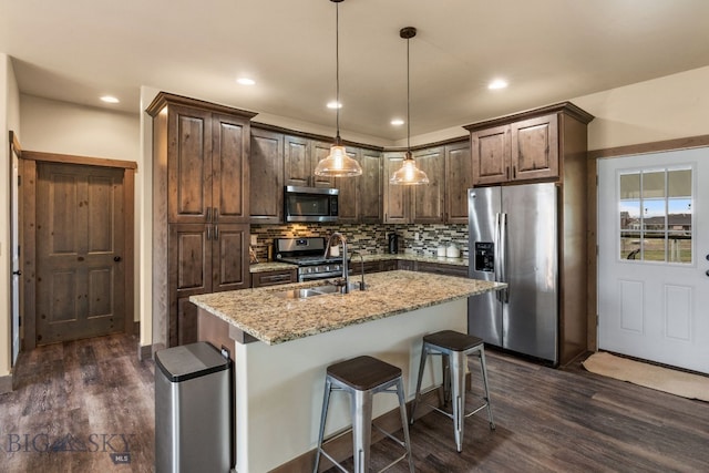 kitchen with light stone countertops, hanging light fixtures, dark hardwood / wood-style floors, a kitchen island with sink, and appliances with stainless steel finishes