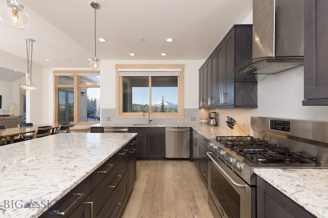 kitchen with light stone counters, stainless steel appliances, wall chimney range hood, pendant lighting, and light hardwood / wood-style flooring