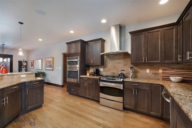 kitchen with dark brown cabinets, hanging light fixtures, light hardwood / wood-style flooring, appliances with stainless steel finishes, and wall chimney range hood