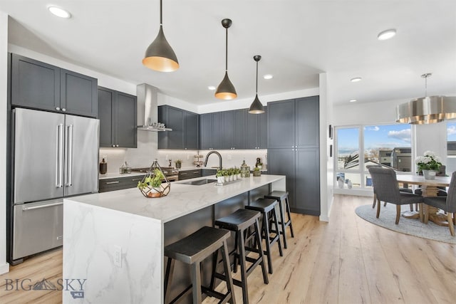 kitchen featuring wall chimney range hood, sink, hanging light fixtures, light wood-type flooring, and stainless steel appliances