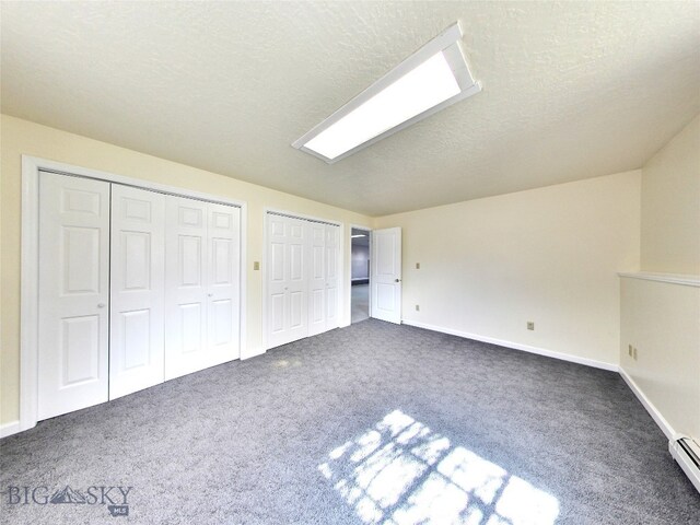 unfurnished bedroom with dark carpet, a textured ceiling, a baseboard heating unit, and multiple closets