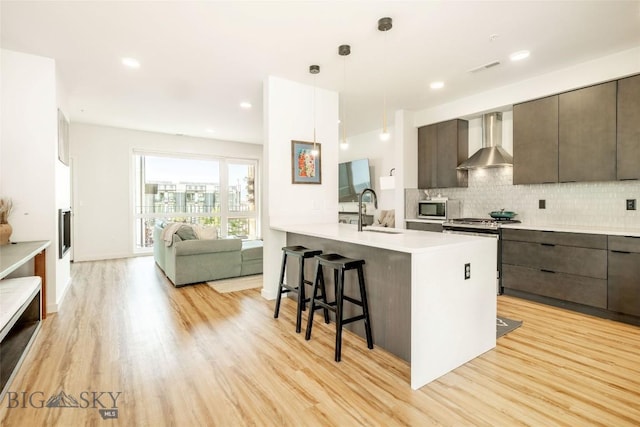 kitchen featuring wall chimney range hood, sink, appliances with stainless steel finishes, light hardwood / wood-style floors, and decorative light fixtures