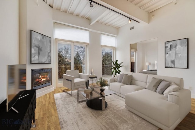 living room with beamed ceiling, track lighting, a towering ceiling, and light hardwood / wood-style flooring