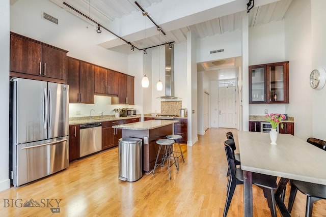 kitchen featuring stainless steel appliances, wall chimney range hood, a kitchen breakfast bar, a towering ceiling, and light hardwood / wood-style floors