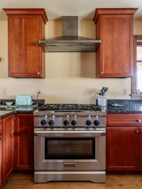 kitchen featuring high end stove, wall chimney range hood, light wood-style flooring, and dark stone counters