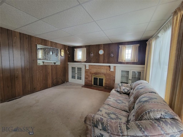 carpeted living room with a paneled ceiling, wooden walls, french doors, and a brick fireplace