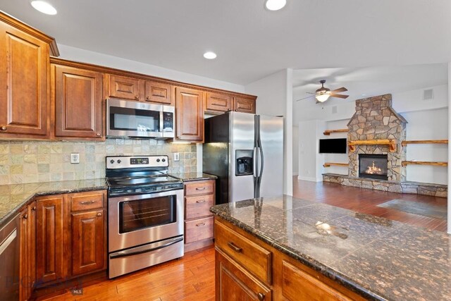 kitchen with light hardwood / wood-style flooring, ceiling fan, appliances with stainless steel finishes, decorative backsplash, and a stone fireplace