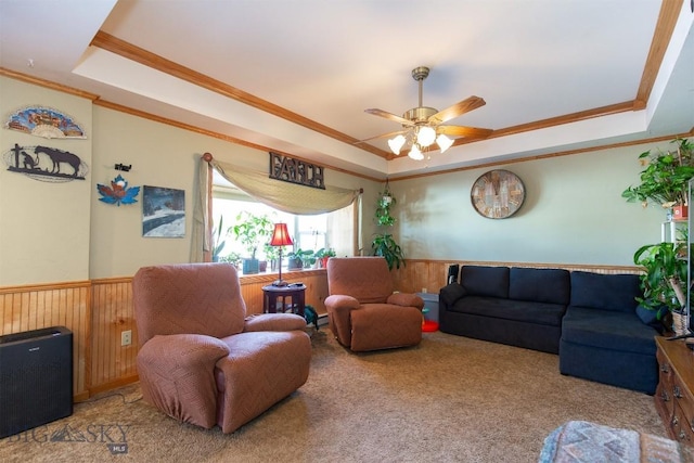 living room with crown molding, a raised ceiling, carpet, a ceiling fan, and wainscoting