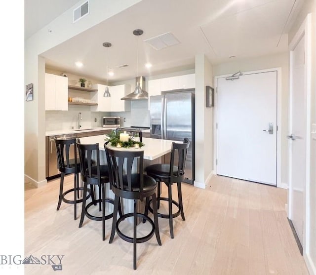 kitchen featuring white cabinetry, stainless steel appliances, backsplash, hanging light fixtures, and wall chimney range hood