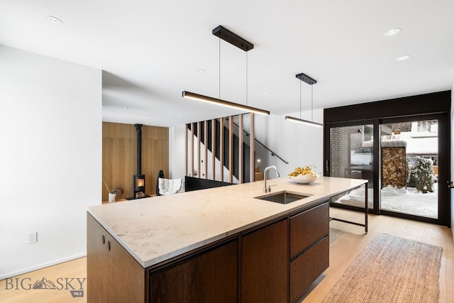 kitchen featuring pendant lighting, an island with sink, sink, a wood stove, and light hardwood / wood-style flooring