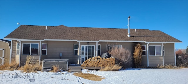 snow covered back of property with a shingled roof and a hot tub