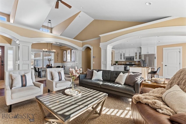 carpeted living room featuring sink, decorative columns, ceiling fan with notable chandelier, and high vaulted ceiling