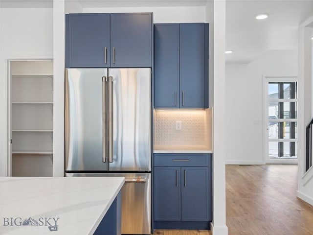 kitchen featuring blue cabinetry, backsplash, light stone counters, high end refrigerator, and light wood-type flooring