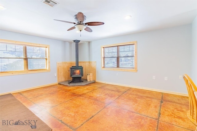 unfurnished living room featuring a ceiling fan, a wood stove, baseboards, and visible vents