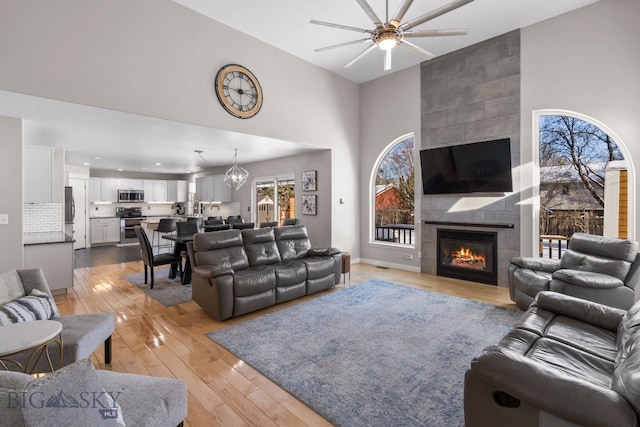 living room featuring a tiled fireplace, ceiling fan with notable chandelier, light hardwood / wood-style flooring, and high vaulted ceiling