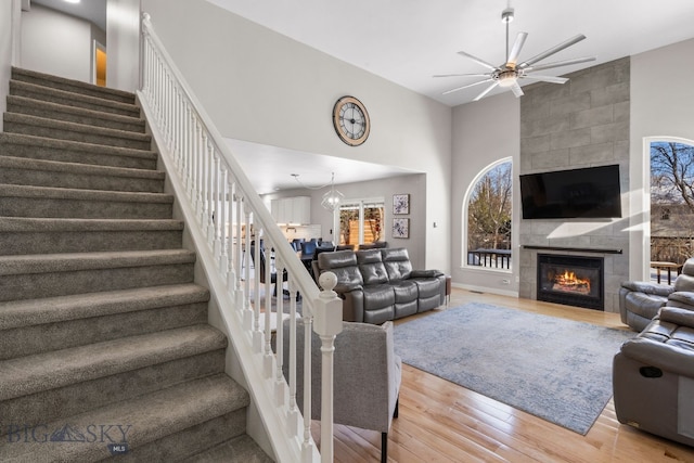 living room featuring wood-type flooring, a tiled fireplace, ceiling fan with notable chandelier, and a high ceiling