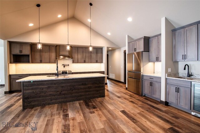 kitchen with an island with sink, sink, stainless steel fridge, hanging light fixtures, and light stone countertops