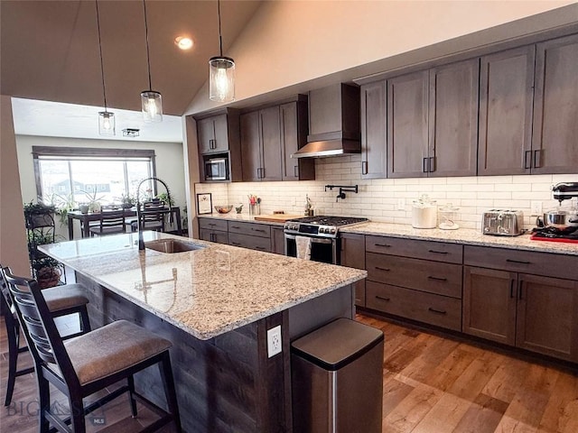 kitchen with a breakfast bar area, wood finished floors, a sink, stainless steel appliances, and wall chimney range hood